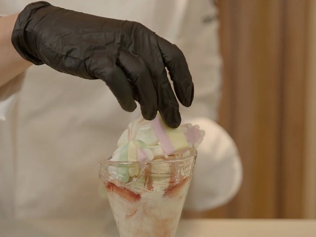 Gloved hand adding a marshmallow to a layered soft serve sundae